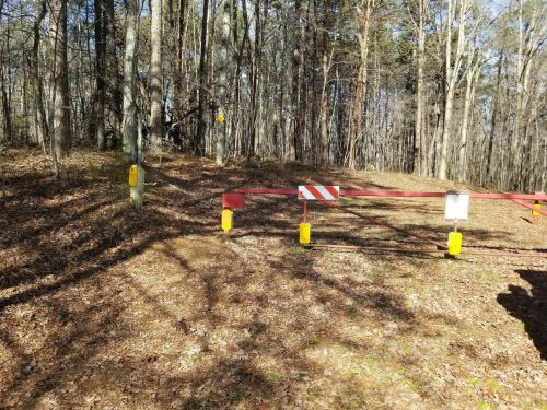 A red barrier gate in a wooded area, surrounded by trees and fallen leaves, indicating restricted access.