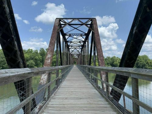 A weathered metal bridge spans a calm river, surrounded by green trees under a blue sky with fluffy clouds.