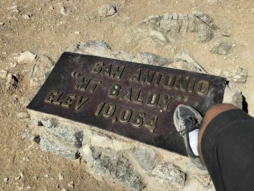 A metal plaque on a rocky surface reads "San Antonio 'Mt Baldy' Elev 10,064," with a person's foot nearby.