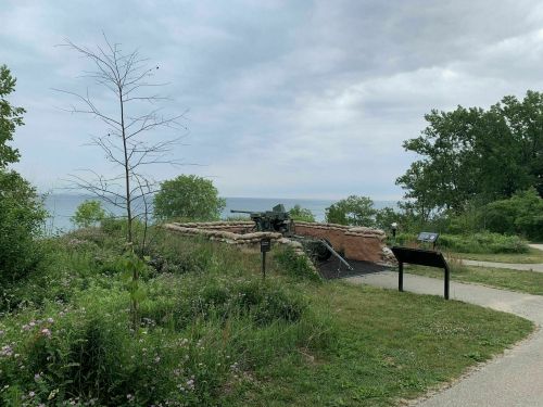 A grassy area with a historical cannon and informational sign, overlooking a calm body of water under a cloudy sky.