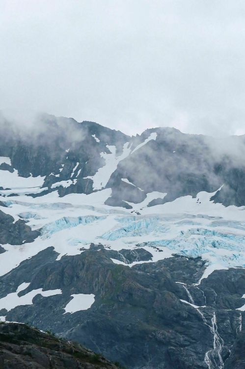 A glacier flows down a rugged mountain landscape, partially obscured by clouds and mist.