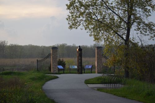 A pathway leads to a gated entrance with informational signs, surrounded by lush greenery and a cloudy sky.