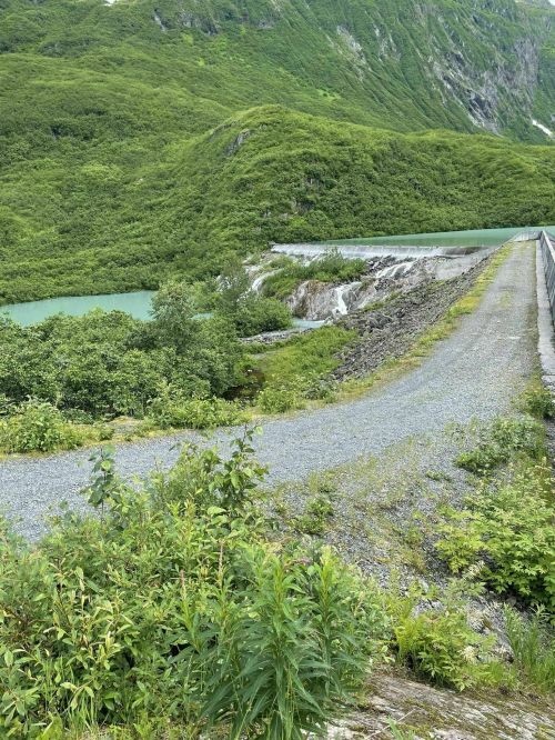 A gravel path leads alongside a river, surrounded by lush green hills and trees under a clear sky.
