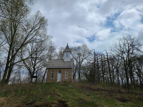 A small, stone church on a hill surrounded by trees under a cloudy sky.