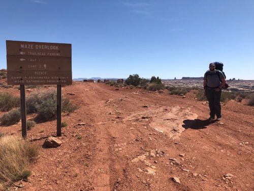 A hiker stands on a dirt path next to a trail sign for Maze Overlook, surrounded by desert landscape.