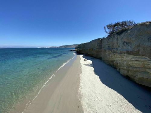 A serene beach scene with clear blue water, sandy shore, and rocky cliffs under a bright blue sky.