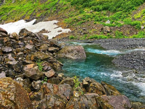 A clear blue stream flows through rocky terrain, surrounded by lush green vegetation and patches of snow.