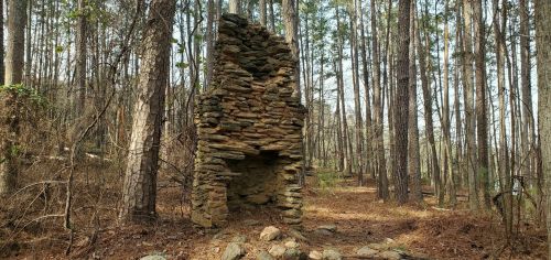 A stone chimney stands among tall trees in a wooded area, surrounded by fallen leaves and rocky ground.
