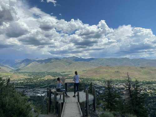 Two people stand on a wooden overlook, gazing at a scenic mountain landscape under a cloudy sky.