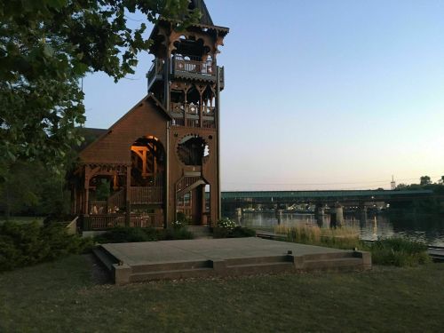 A wooden structure by a river at dusk, with a bridge in the background and greenery surrounding the area.