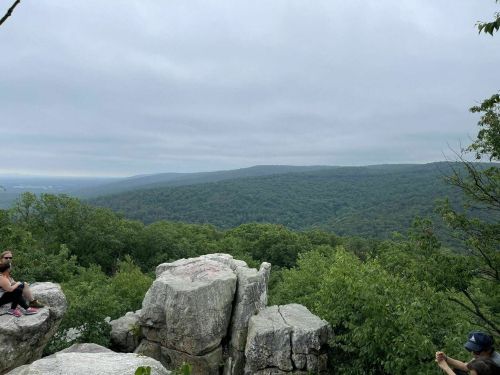 A scenic view from a rocky outcrop, overlooking a lush green valley under a cloudy sky. Two people are visible.