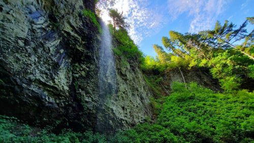 A lush green landscape with a waterfall cascading down a rocky cliff under a bright blue sky.