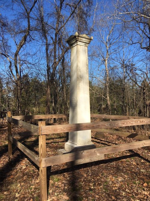 A tall stone pillar surrounded by a wooden fence, set in a wooded area with bare trees and a clear blue sky.