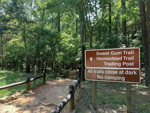 Trail sign in a wooded area, indicating Sweet Gum Trail and Homestead Trail, with a note about closing at dark.