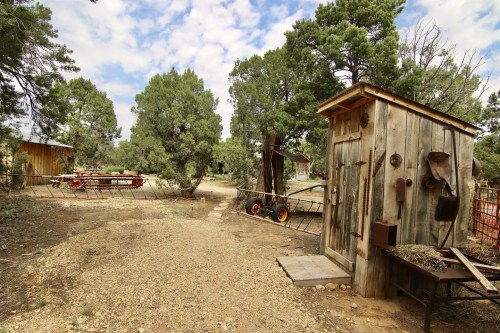 A rustic wooden shed stands beside a gravel path, surrounded by trees and farm equipment in a rural setting.