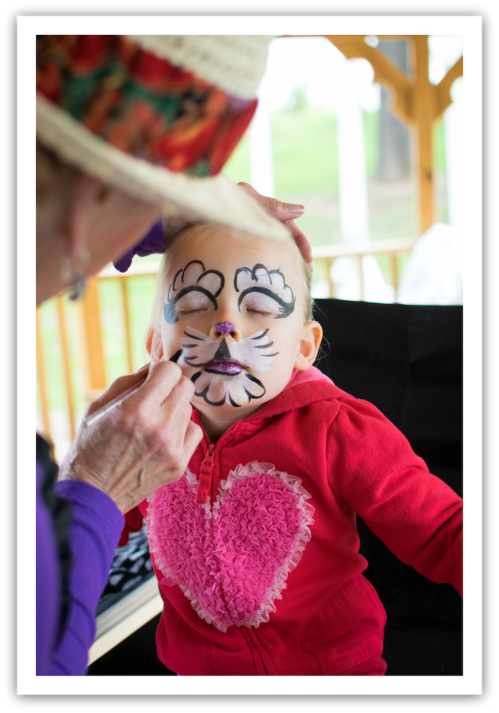 A child with face paint resembling a cat sits quietly while an adult applies more paint. The child wears a heart-patterned hoodie.