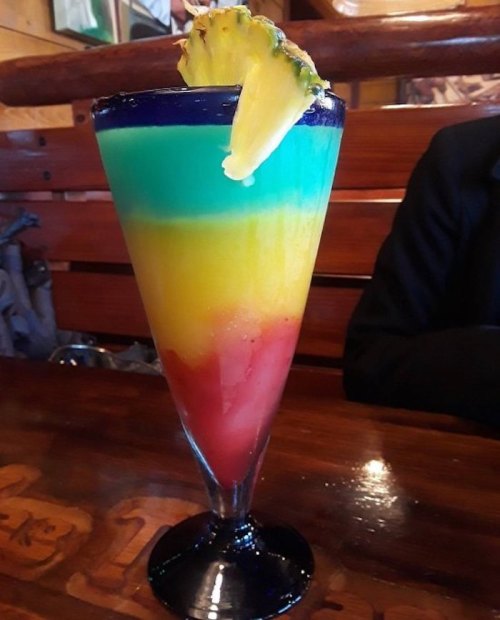 A colorful layered drink in a tall glass, featuring red, yellow, green, and blue hues, garnished with a pineapple slice.