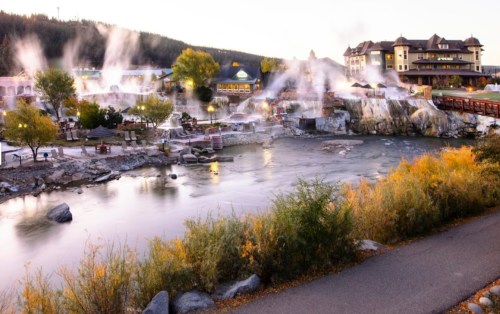 A scenic view of a hot springs resort with steam rising, surrounded by trees and a river, under a clear sky.