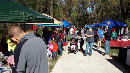 A bustling outdoor market with colorful tents, vendors, and families walking along a path on a sunny day.