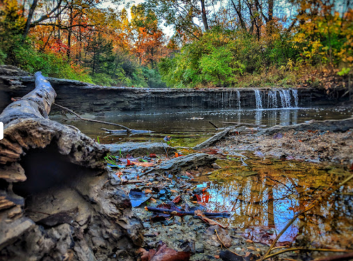 A serene landscape featuring a small waterfall, autumn foliage, and a calm stream reflecting the colorful trees.