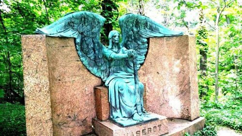 A bronze angel statue with wings, seated on a stone pedestal, surrounded by lush greenery.