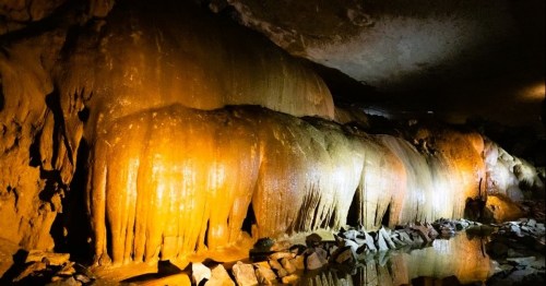 Stalactites and rock formations illuminated in a cave, reflecting in a shallow pool of water below.
