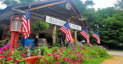 A rustic store with a sign reading "Old-Sale Store," adorned with American flags and surrounded by colorful flowers.