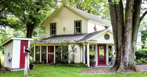A charming two-story house surrounded by trees, featuring a red door and a small shed nearby.