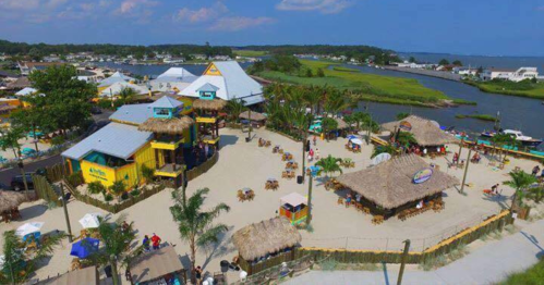 Aerial view of a vibrant beachside restaurant area with palm trees, sandy spaces, and water in the background.