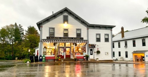 A quaint general store with a white facade, surrounded by trees, on a rainy day. Warm lights glow from the entrance.