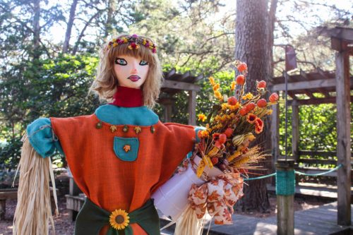 A colorful scarecrow with a flower crown holds a bouquet of autumn flowers in a garden setting.
