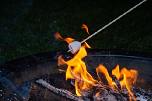A marshmallow on a stick is being roasted over a campfire, with flames and glowing embers in the background.