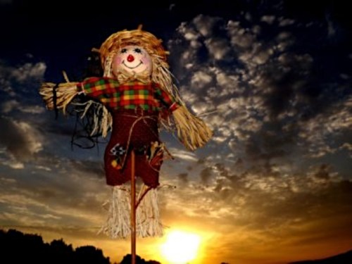 A colorful scarecrow stands against a dramatic sunset with clouds, silhouetted in the foreground.