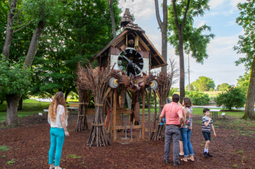 A whimsical structure resembling a house made of natural materials, with people exploring it in a green park setting.