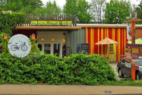 Colorful storefront of a market with outdoor seating and greenery in front. Signs indicate various offerings.