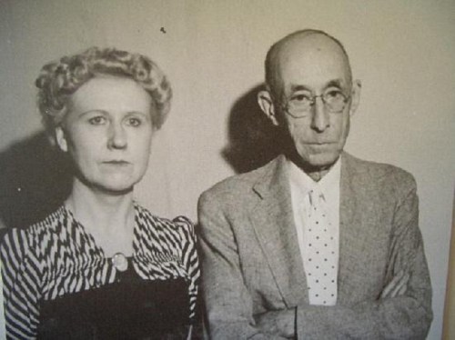 A black-and-white photo of a serious man and woman standing side by side, both dressed in formal attire.