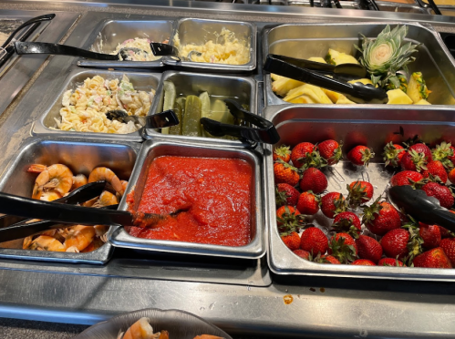 A buffet spread featuring pasta salad, pickles, shrimp, marinara sauce, pineapple, and fresh strawberries.