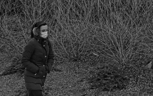 A person wearing a mask and winter coat walks through a barren landscape with sparse vegetation.