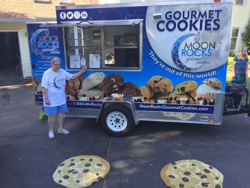 A woman stands next to a food truck named "Moon Rocks Gourmet Cookies," featuring large cookie designs on the ground.