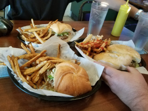 Three plates of burgers with fries on a wooden table, one hand reaching for a sandwich.