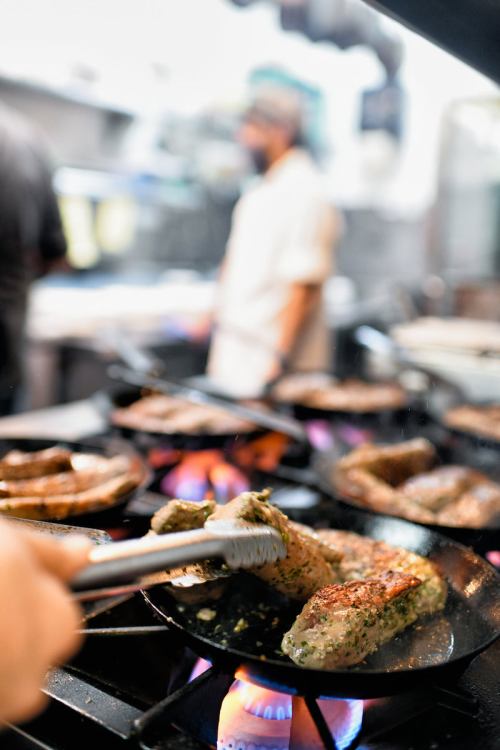A chef flips marinated meat on a sizzling grill, with flames and a blurred kitchen background.