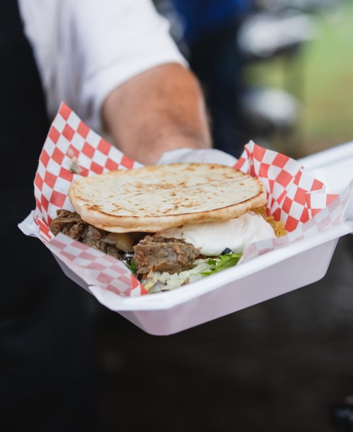 A person holds a tray with a flatbread, meat, rice, and vegetables, all served in a checkered paper-lined container.