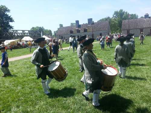 A group of drummers in historical attire perform on a grassy field during a lively outdoor event.