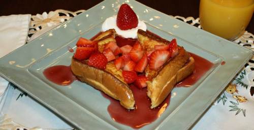 A plate of French toast topped with strawberries and whipped cream, drizzled with syrup, alongside a glass of orange juice.