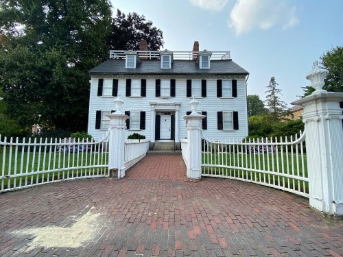 A white colonial-style house with black shutters, surrounded by a white picket fence and a brick pathway.
