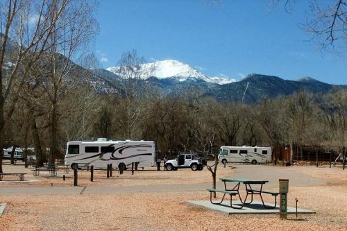 A scenic RV park with two RVs, picnic tables, and snow-capped mountains in the background under a clear blue sky.