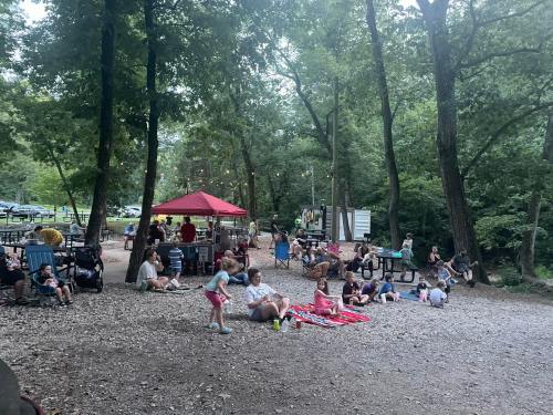 A gathering of people sitting on the ground and at picnic tables in a wooded area, enjoying a sunny day outdoors.
