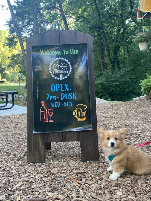 A corgi sits beside a sign that reads "Welcome to the Beer Garden, Open 2 PM - Dusk, Wed - Sun" in a wooded area.