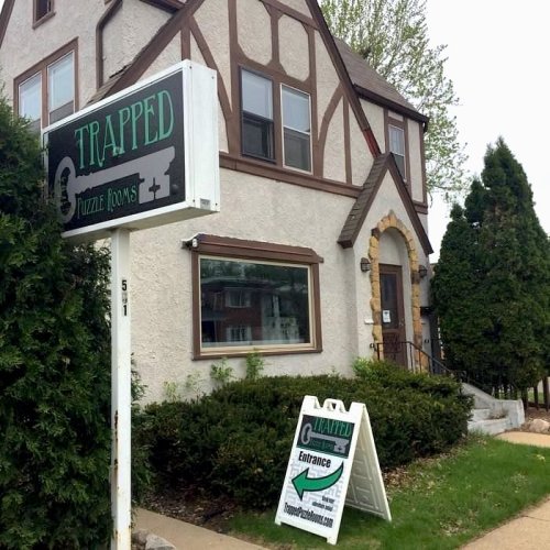 A building with a sign reading "Trapped" and a sandwich board advertising escape rooms, surrounded by greenery.