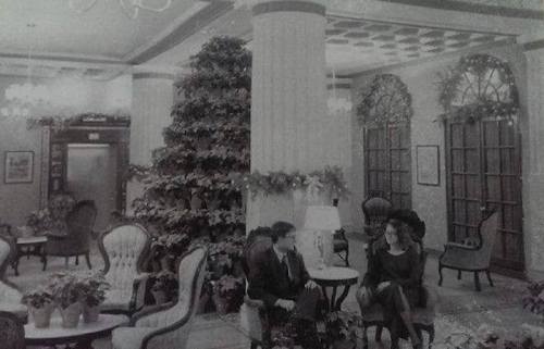 A vintage hotel lobby with elegant seating, a large Christmas tree, and two people engaged in conversation.